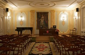 Interior del auditorio del museo. En el escenario se encuentran, a la izquierda, un piano de cola, y en el centro una mesa y una retrato de una reina. 