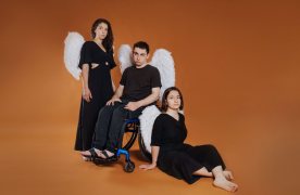Dues noies i un noi vestits de negre amb ales d'àngel a l'esquena. El noi, enmig de les dues, assegut en una cadira de rodes.