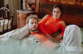 Un noi i una noia estirats a un llit mirant a càmera. Van vestits de blanc i taronja