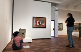 Una persona aseguda al terra devant d'un quadre observa mentre que un altre persona el mira també