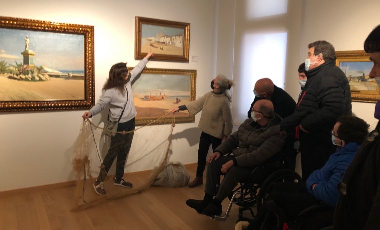 La guia explica a un grup de persones, algunes amb cadires de rodes, un quadre on es pot veure unes xarxes de pescadors com les que mostra al públic. 