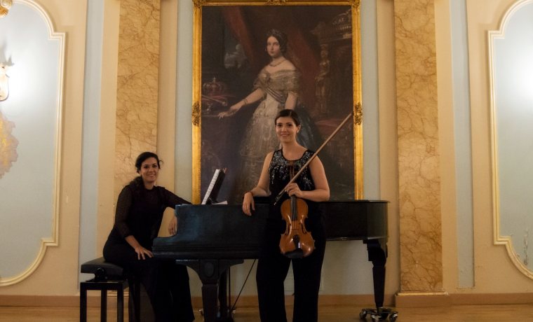 Fotografía en la que aparece una mujer joven sentada en posición de tocar un piano de colar y, delante del piano, una violinista joven con su violín. Ambas visten de negro