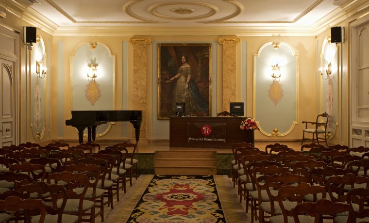 Fotografía general del auditorio del museo. En el fondo se ve el escenario con un piano a la izquierda y una mesa grande con un portátil a la derecha.