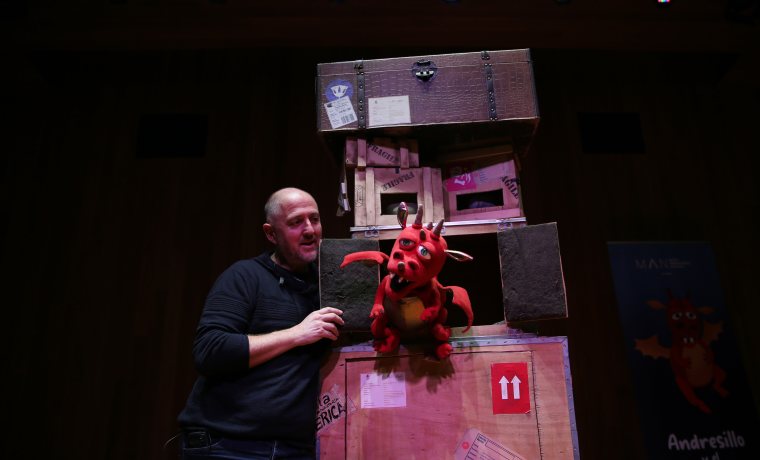 Decorado de teatral con cajas marrones con pegatinas, actor de negro, y marioneta de dragón rojo