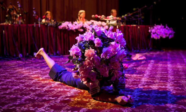Fotografia de l'obra. Es veu una actriu estirada a terra amb una màscara feta de roses